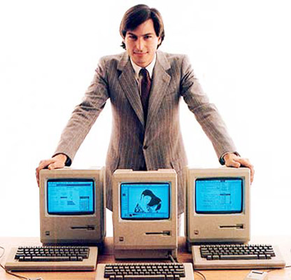 A vintage photo of Steve Jobs standing in front of three early Mac computers.